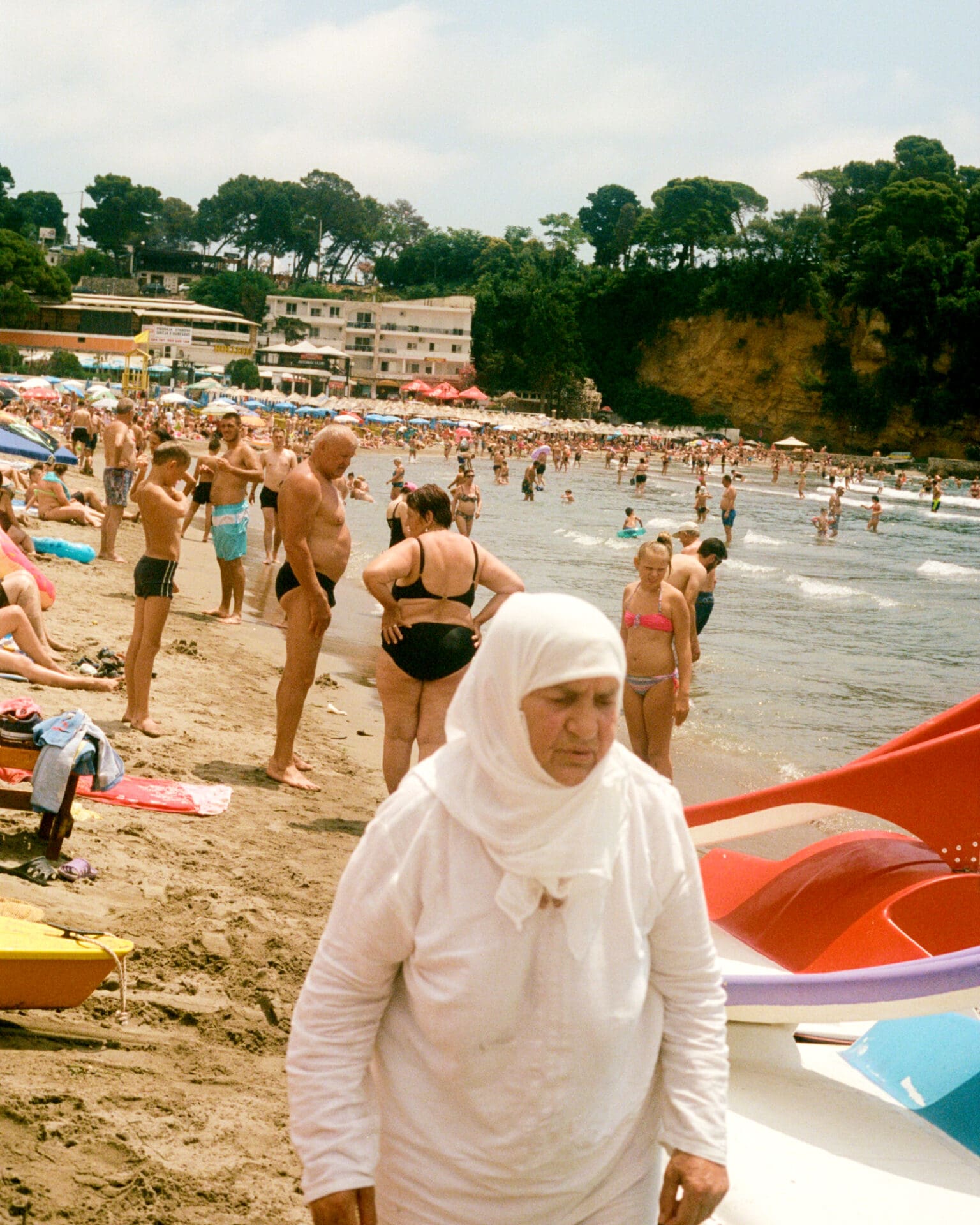Hazel Gaskin photographs Albania | a woman in a white headscarf and robe on the beach, with other beachgoers behind her
