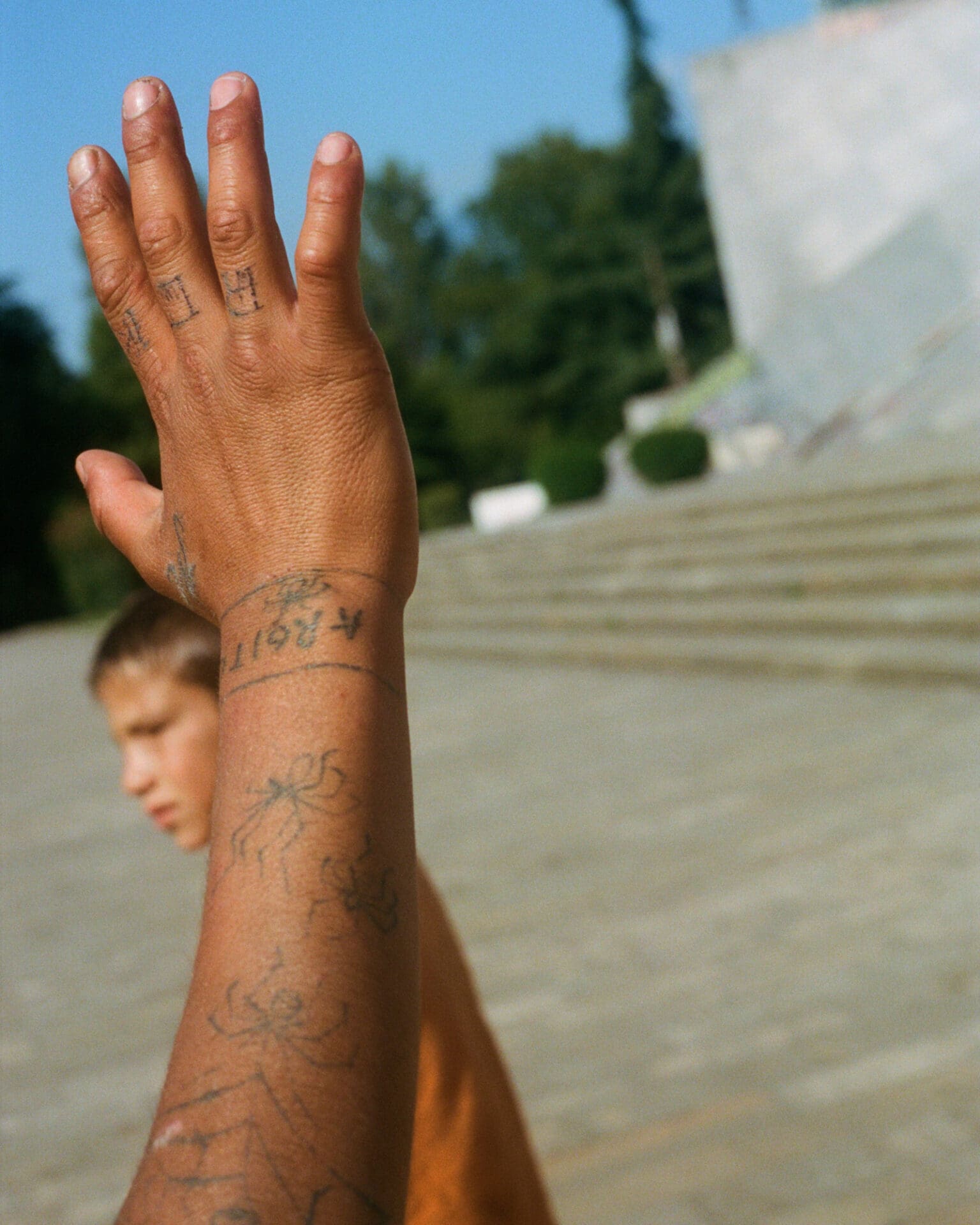 Hazel Gaskin photographs Albania | A boy raises his arm, tattooed with spiders, against a concrete backdrop