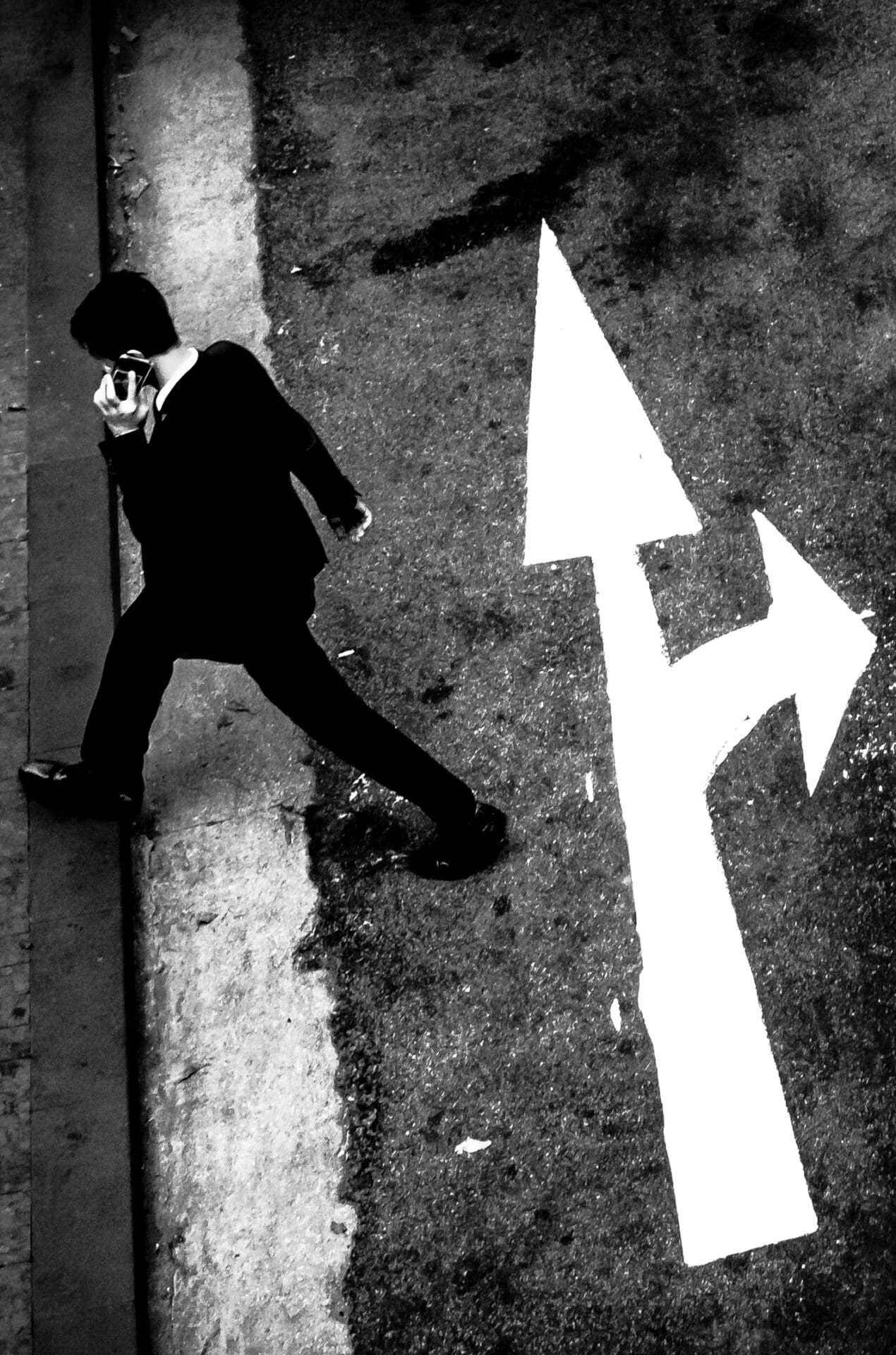Jason Au | A dark-suited man on his phone crosses the road at the business heart of Hong Kong. His mid-stride motion juxtaposes with the white arrows marking on the road