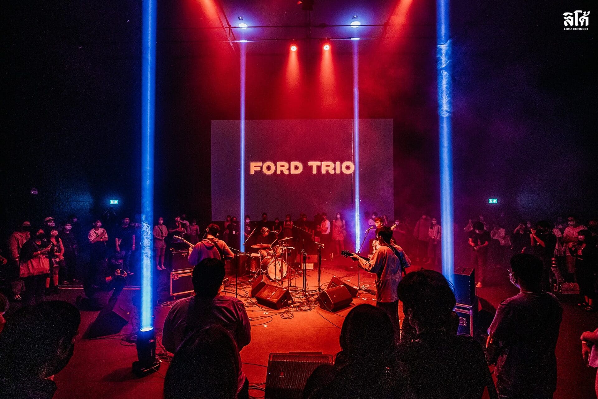 The best museums and galleries in Bangkok | a view inside the performance space of Lido Connect, where three guitarists and a drummer perform in the round, surrounded by an audience. A projection on the back of the wall says Ford Trio, and four lights create columns of blue light against a moody red ambiance.
