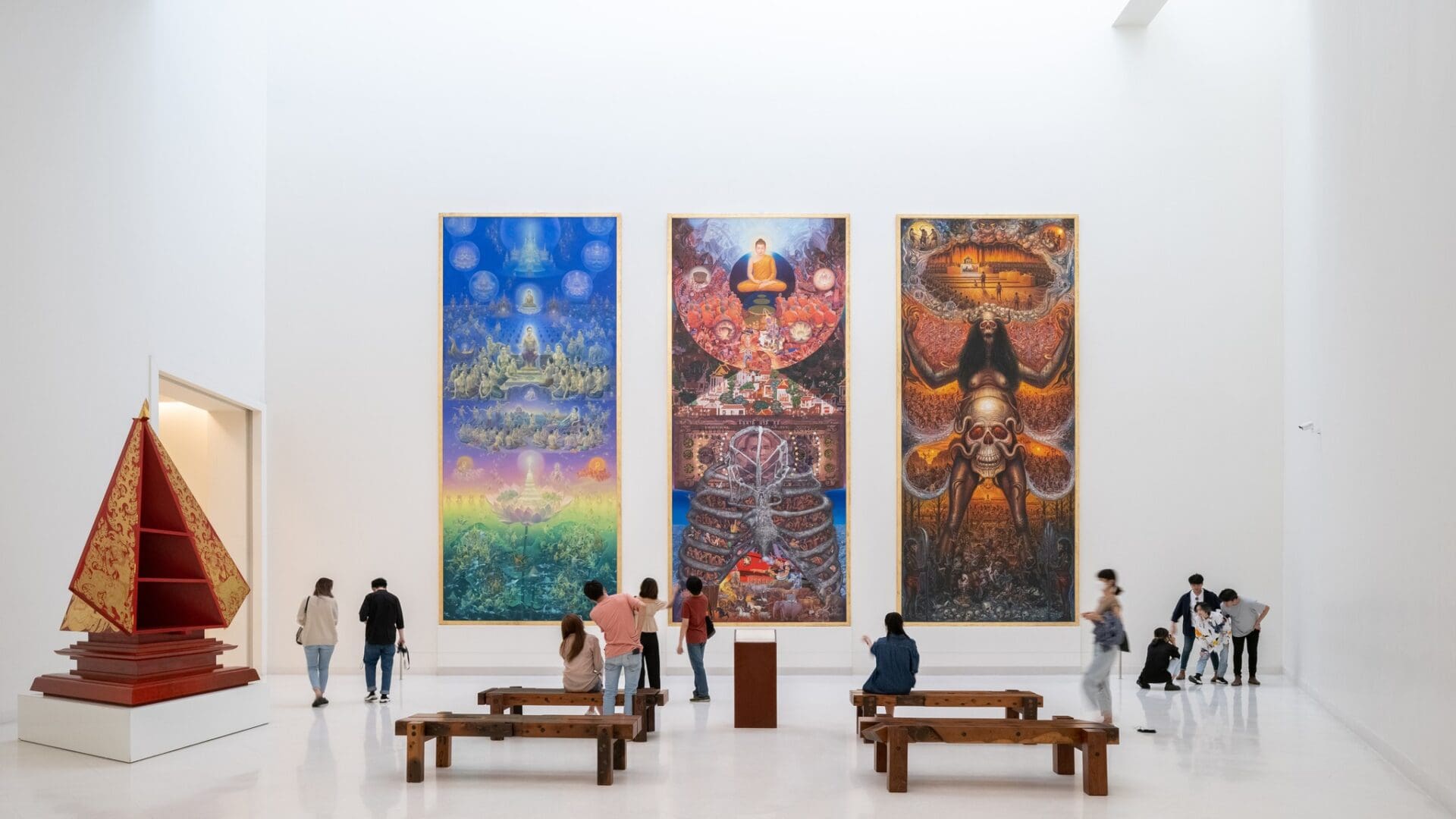 The best museums and galleries in Bangkok | a view of a tall white walled gallery inside Moca Bangkok, with three long colourful canvases arranged next to one another on the far wall. People gather among benches to view the art works