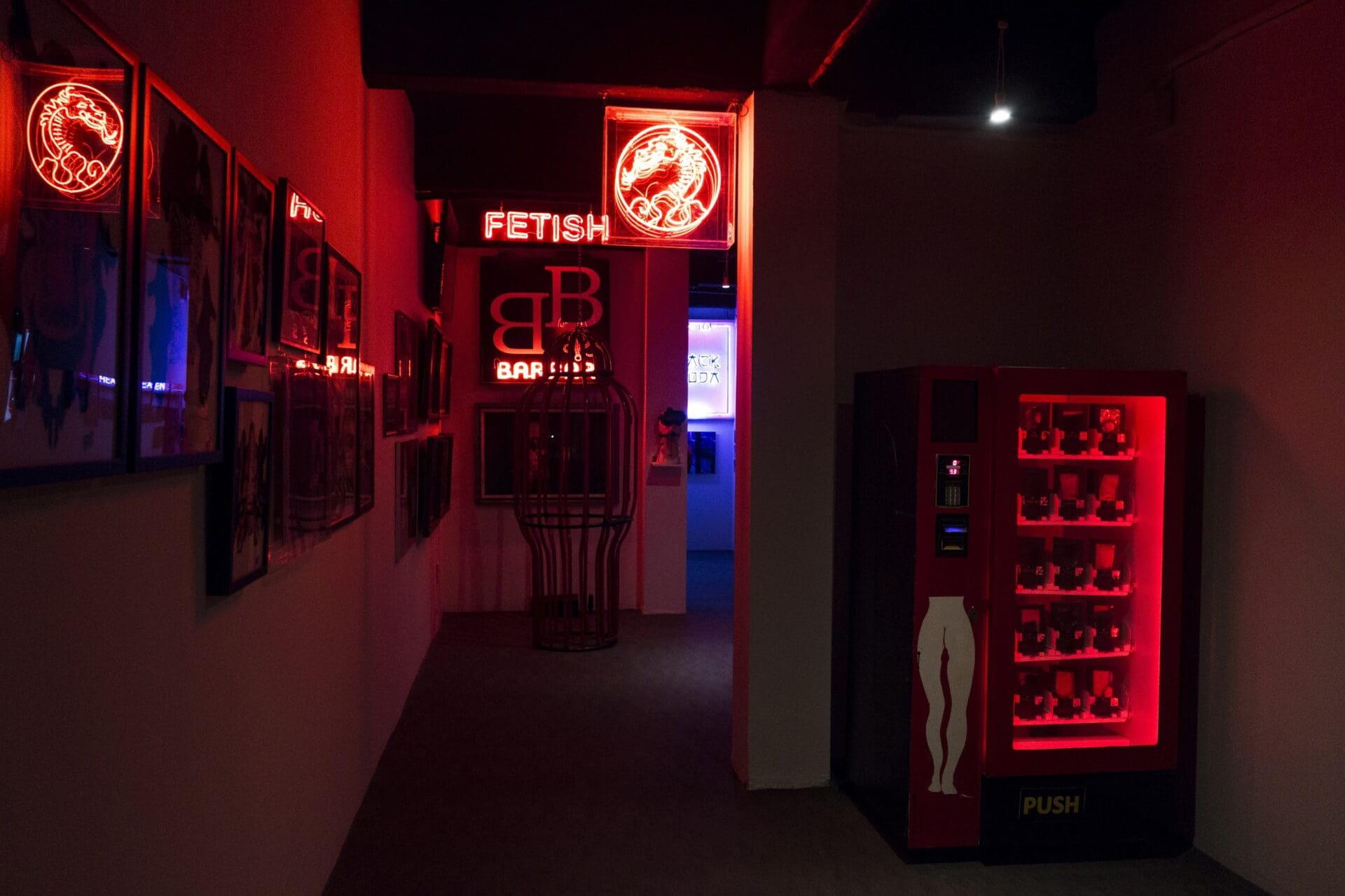The best museums and galleries in Bangkok | a view inside Patpong Museum, with lots of shadows and red neon lighting. A neon dragon sign is visible, as is a vending machine, a cage, and the word FETISH.