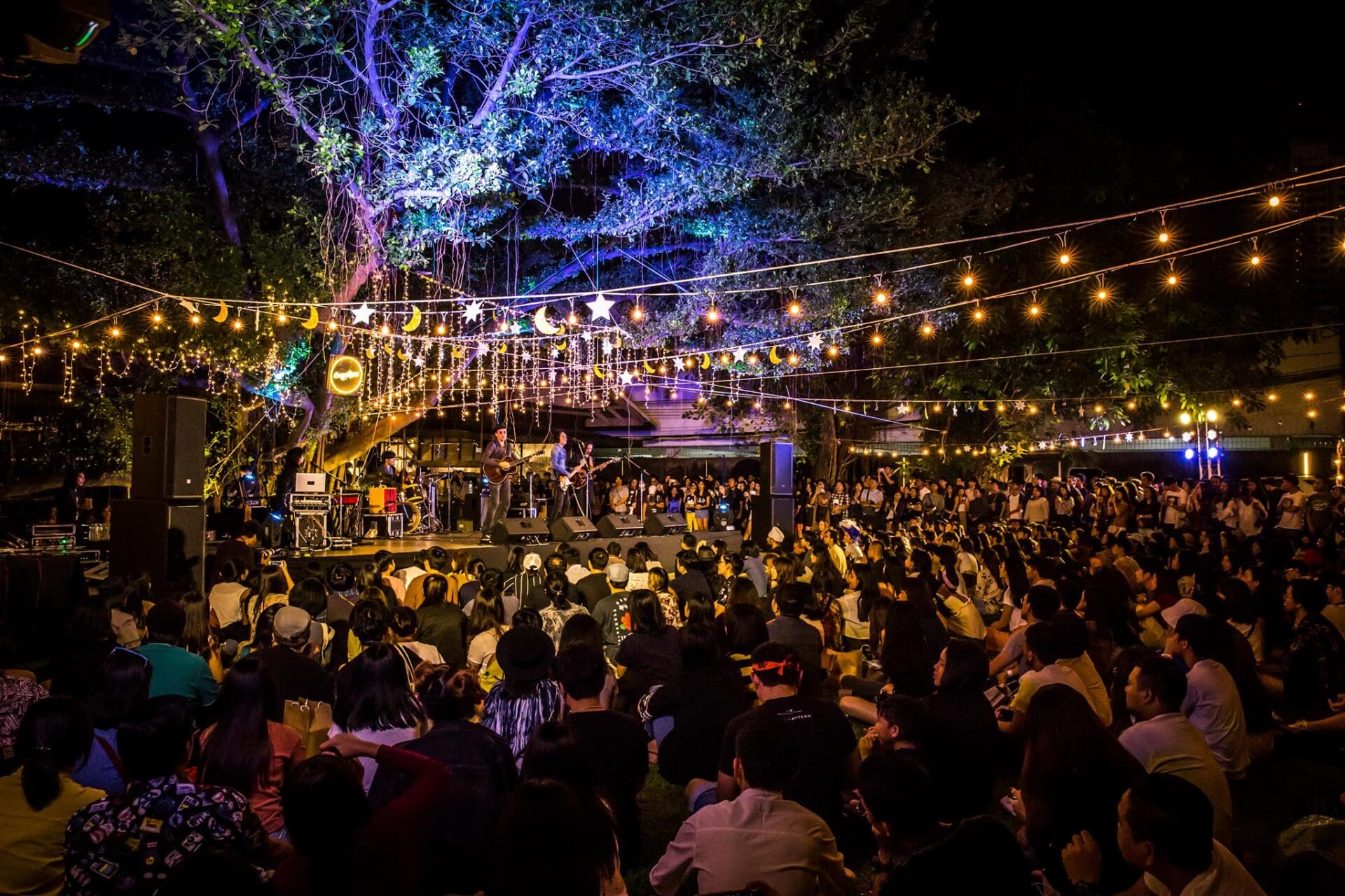 The best galleries and museums in Bangkok | a view of a big crowd, sat cross-legged on the floor, watching a band play on a stage below a tree at night. The tree is lit up from below, and rows of bulbs run from the tree and across the audience
