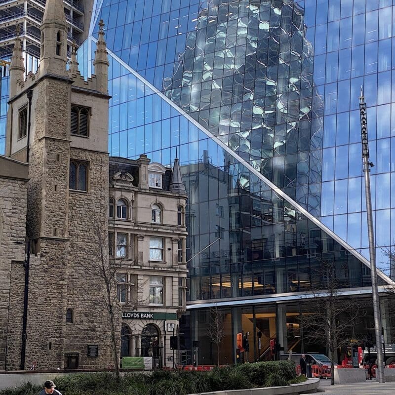 Local's guide to London | A grey brick church stands in the shadow of a mirrored building in the City of London, with the Gherkin reflected in its windows