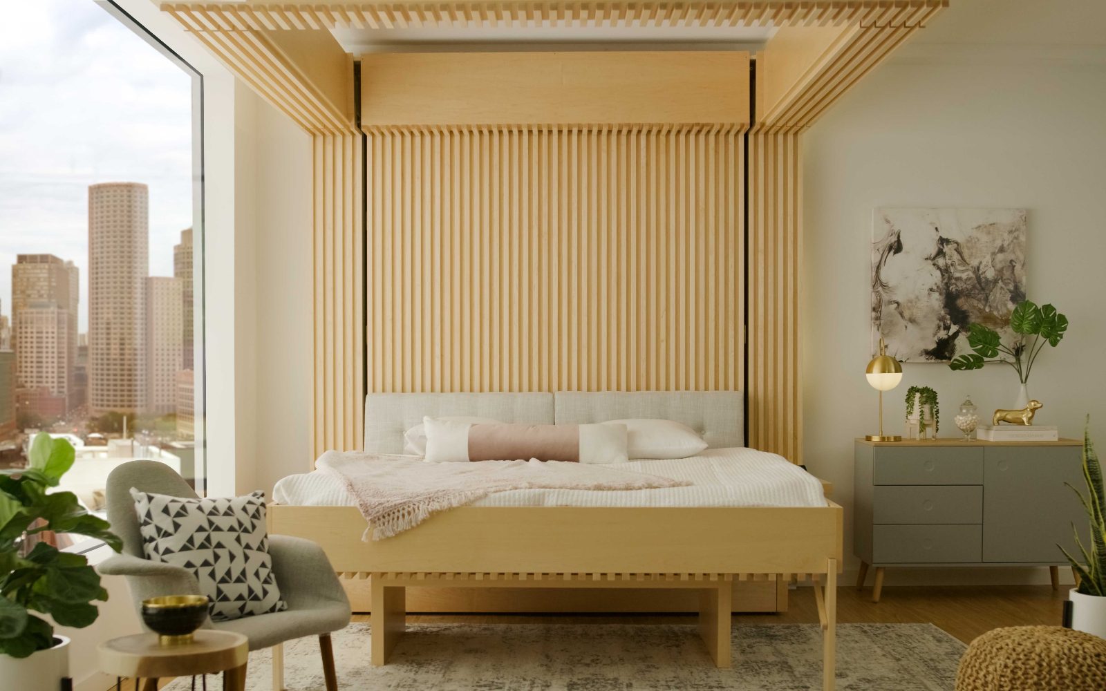 Hotel and digital nomads | The futon-style Ori cloud folding bed, with a light-wood headboard, in a guest room with a bedside table, chair and plants and city views from the floor-to-ceiling window
