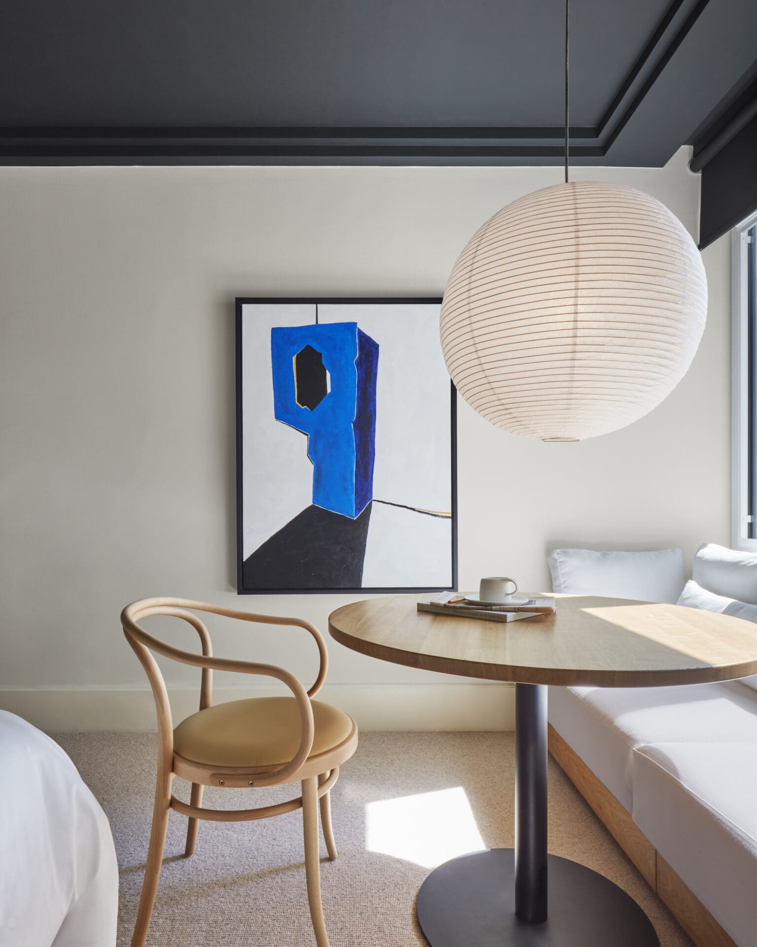 Best hotels in Shoreditch, Dalston and Hackney | A view of a bedroom in One Hundred Shoreditch. A paper light lantern hangs from the ceiling over a round table, where a wooden cane seat and a sofa can be seen. A painting hangs on the wall. The ceiling is painted in a dark grey, and the walls a light neutral shade.