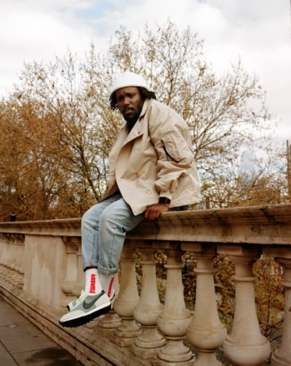 Akwasi Brenya-Mensa | Akwasi wearing jeans, white trainers, a white beanie and a trench coat sat on a rail at Somerset House
