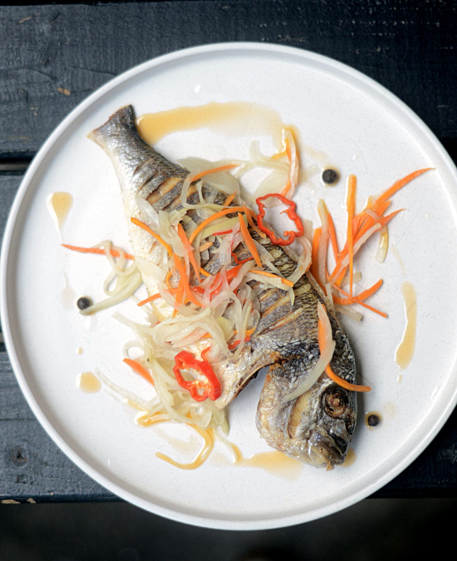 London's Caribbean community | A grilled fish lying on its side on a round white plate, scattered with thinly sliced chopping vegetables like carrots and chillies