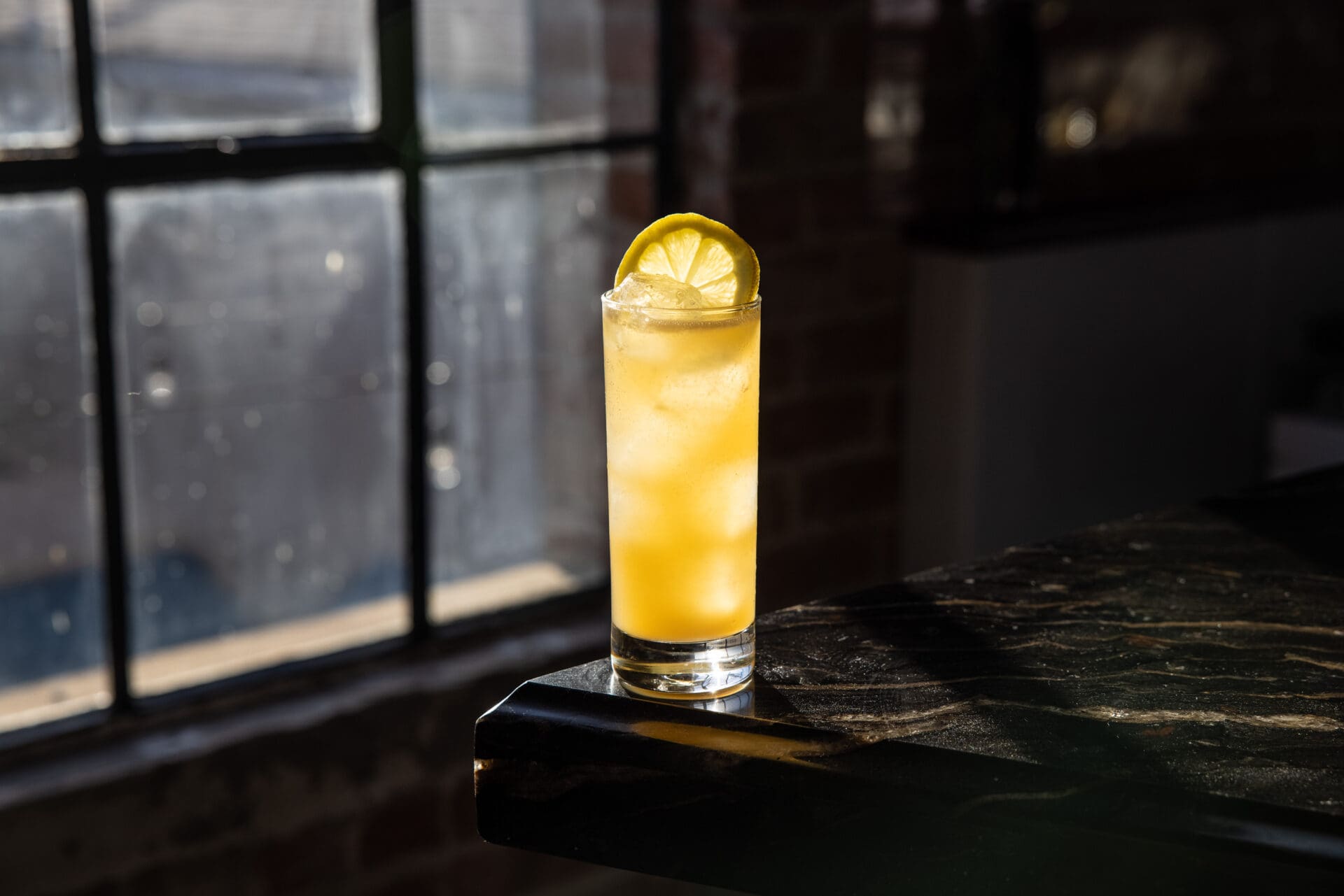 Best rooftop bars LA | A yellow cocktail in a tall glass with a lemon slice for a garnish, silhouetted against a warehouse-style window