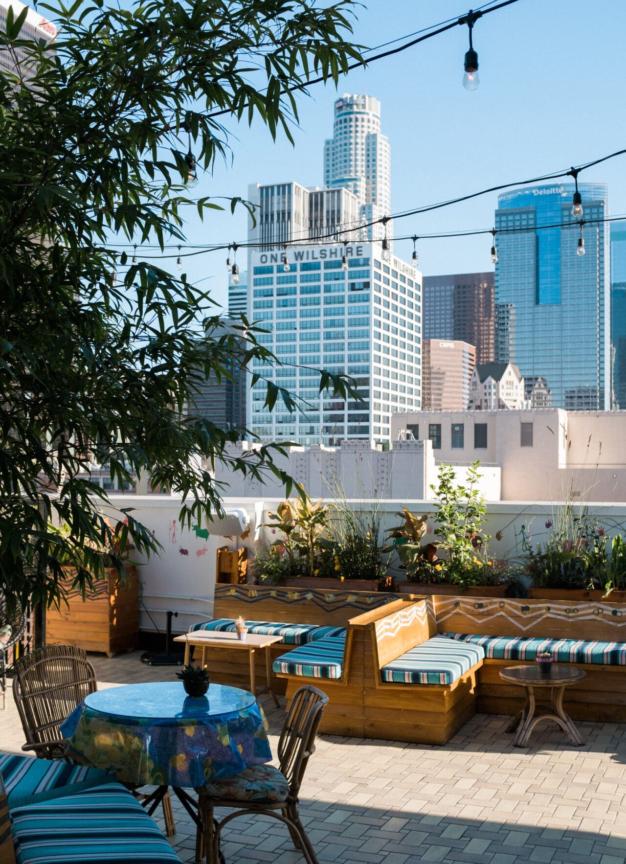 Best rooftop bars in LA | a tree, some wooden bench seating with blue striped cushions and tables at rooftop bar the Broken Shaker at the Freehand Hotel, with a backdrop of blue sky and skyscrapers