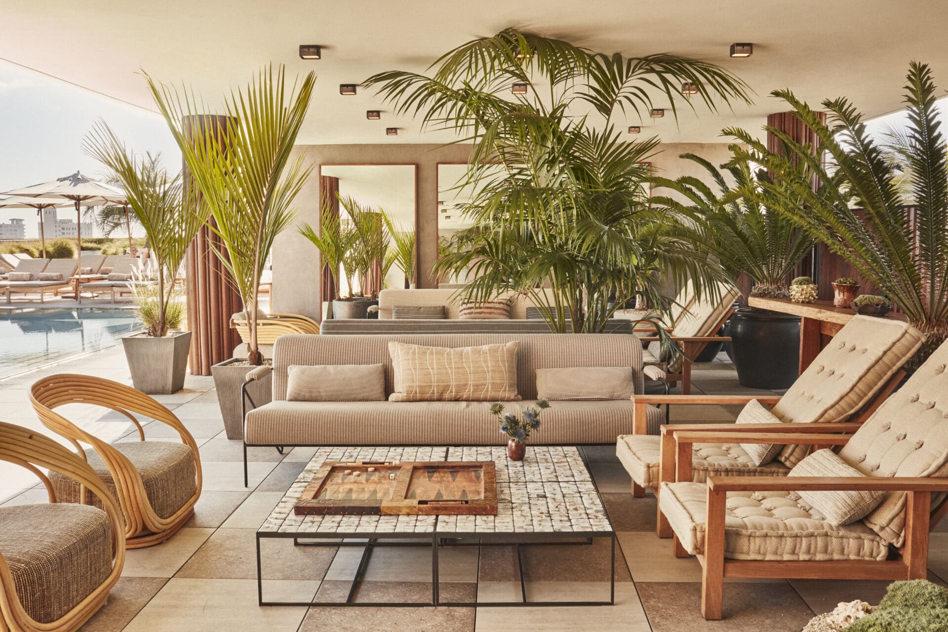 Sleek white interiors with palm trees overlooking the pool at the Santa Monic Proper Hotle's Calabra bar; with a low table topped by a backgammon set and reclining wooden chairs