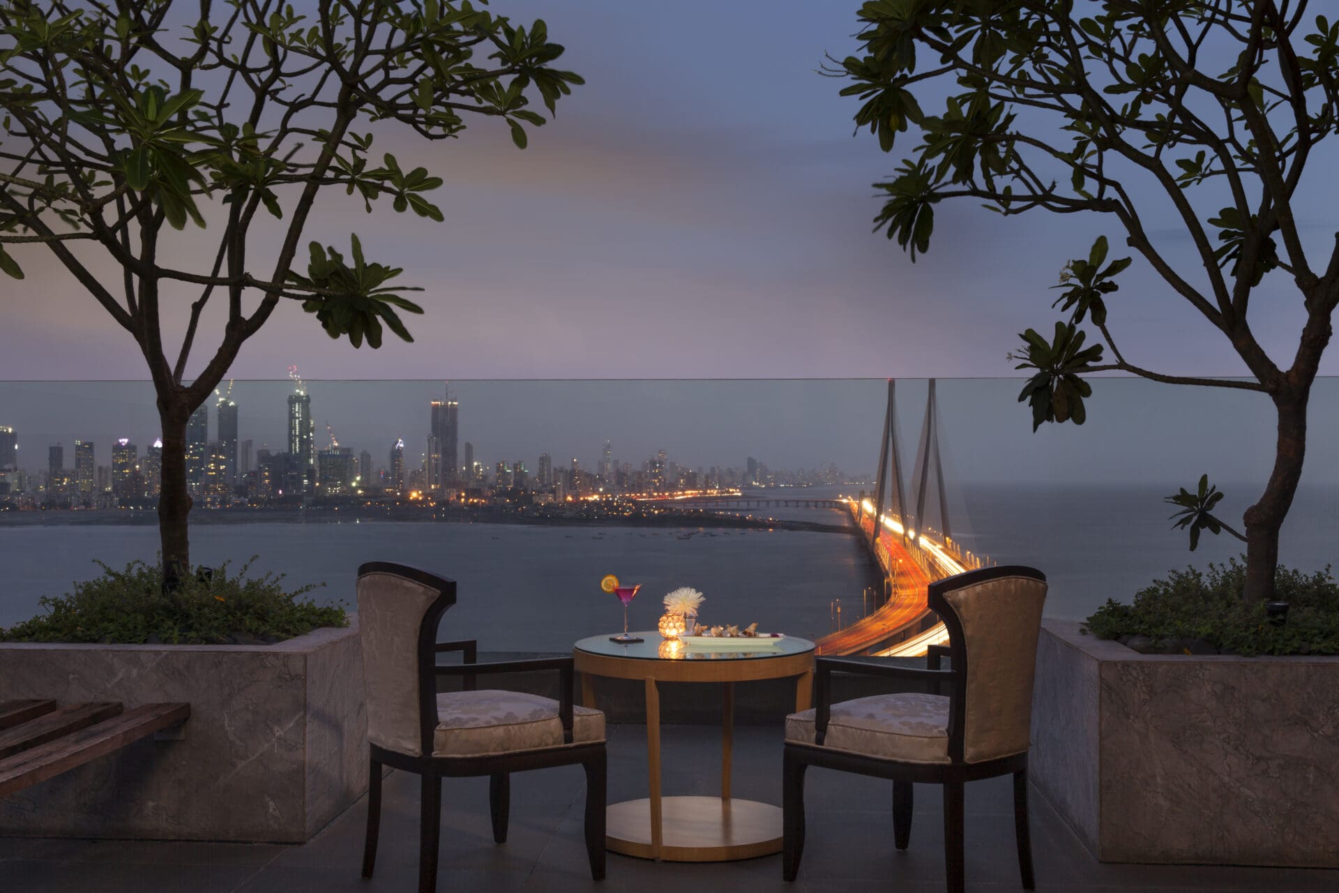 The best hotels in Mumbai | A view of the Sealink at dusk from the terrace at Taj Lands End, with a table and two chairs and two potted trees in the foreground.