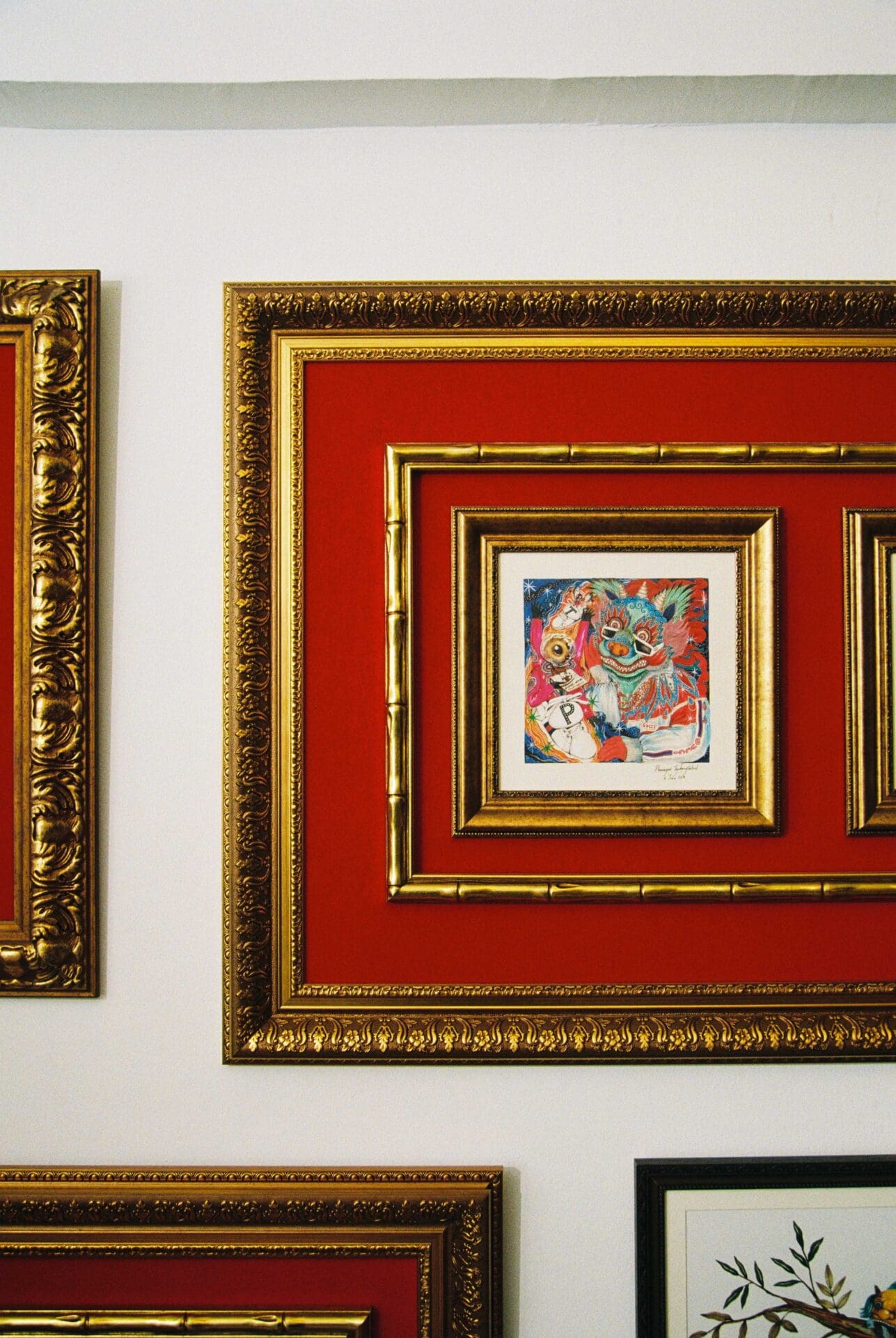 Phannapast | A small, square colourful painting sits on a red background inside a gold-edged frame, hung on a white wall