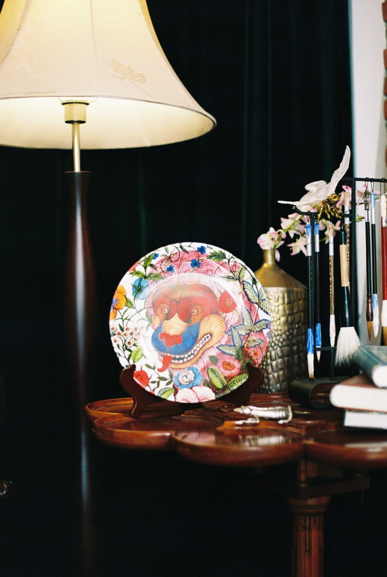 Phannapast | A multi-coloured patterned china plate is displayed on a dark wood table, in front of a darkly coloured wall and next to an vintage-style floor lamp with large lampshade