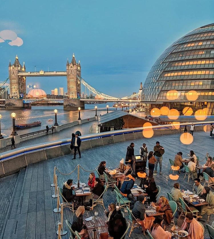 Best outdoor restaurants London | A view over the river Thames and people dining outside under globe-shaped lights at dusk, in front of the lit-up Tower Bridge