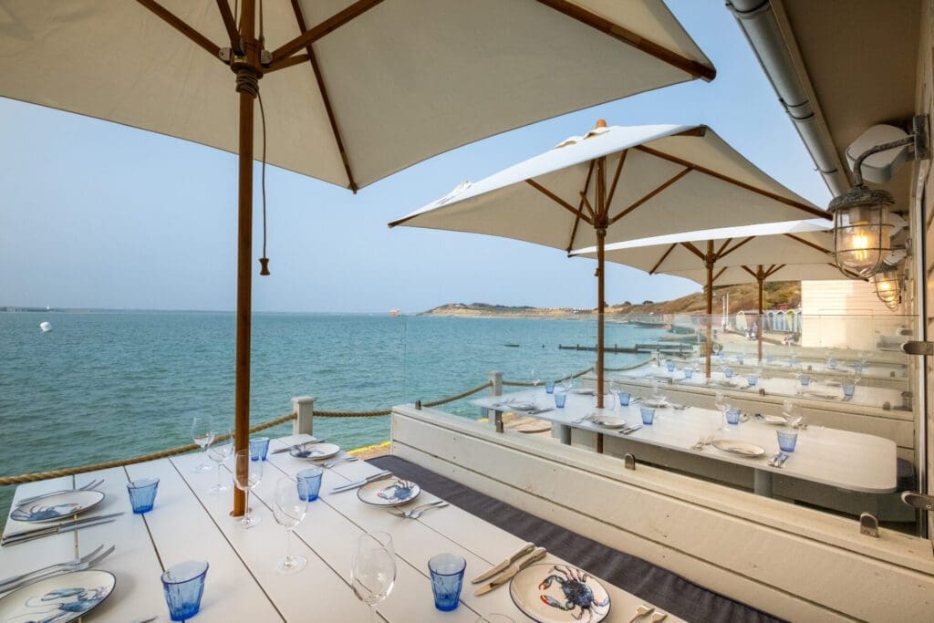 Isle of Wight | white wooden benches and tables are laid out with cutlery, blue glassware and plates painted with blue crabs, under white umbrellas and by the sea at The Hut