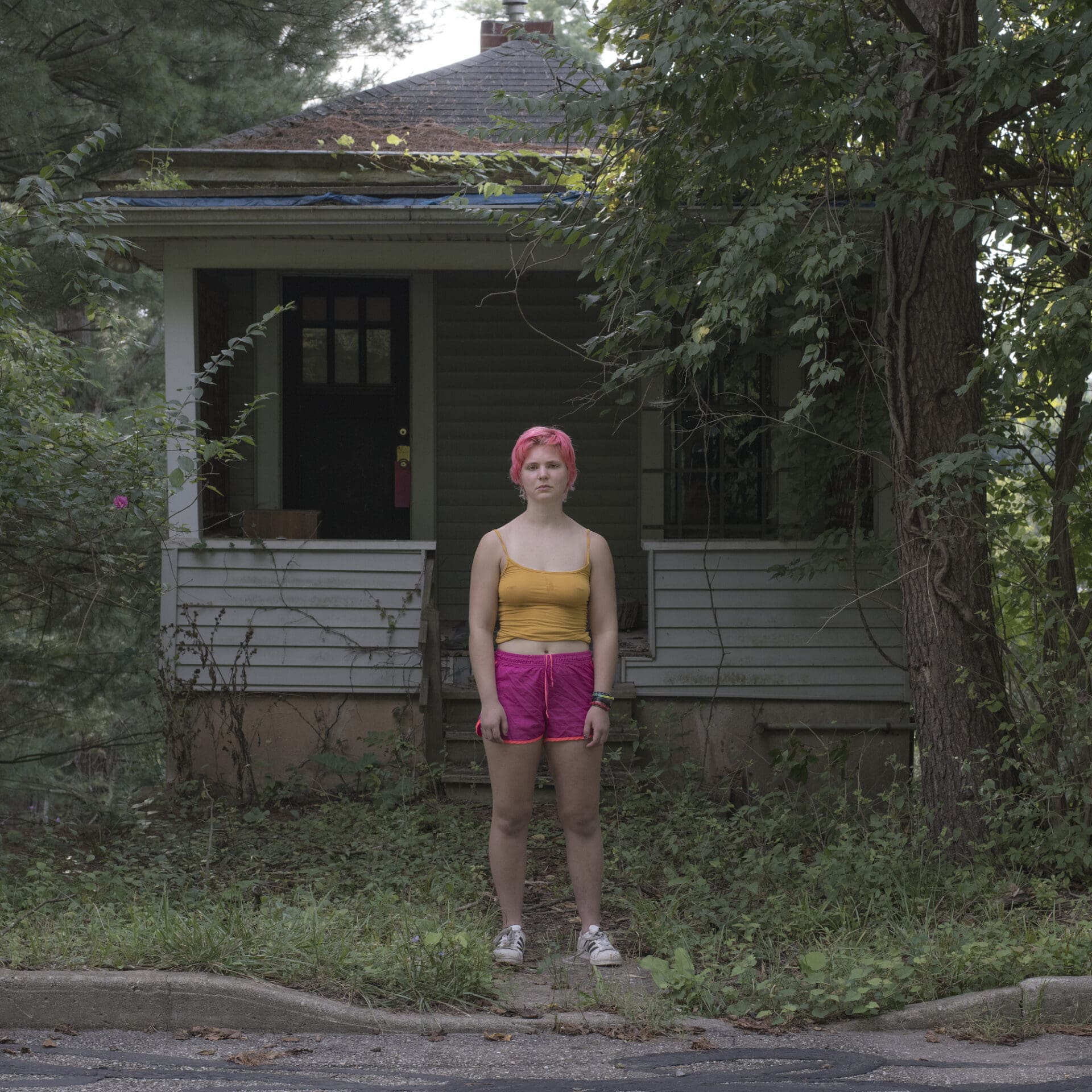 Photographer Rich-Joseph Facun | A woman with short pink hair wearing a yellow vest top and bright-pink shorts stands in front of a white-boarded single-storey house