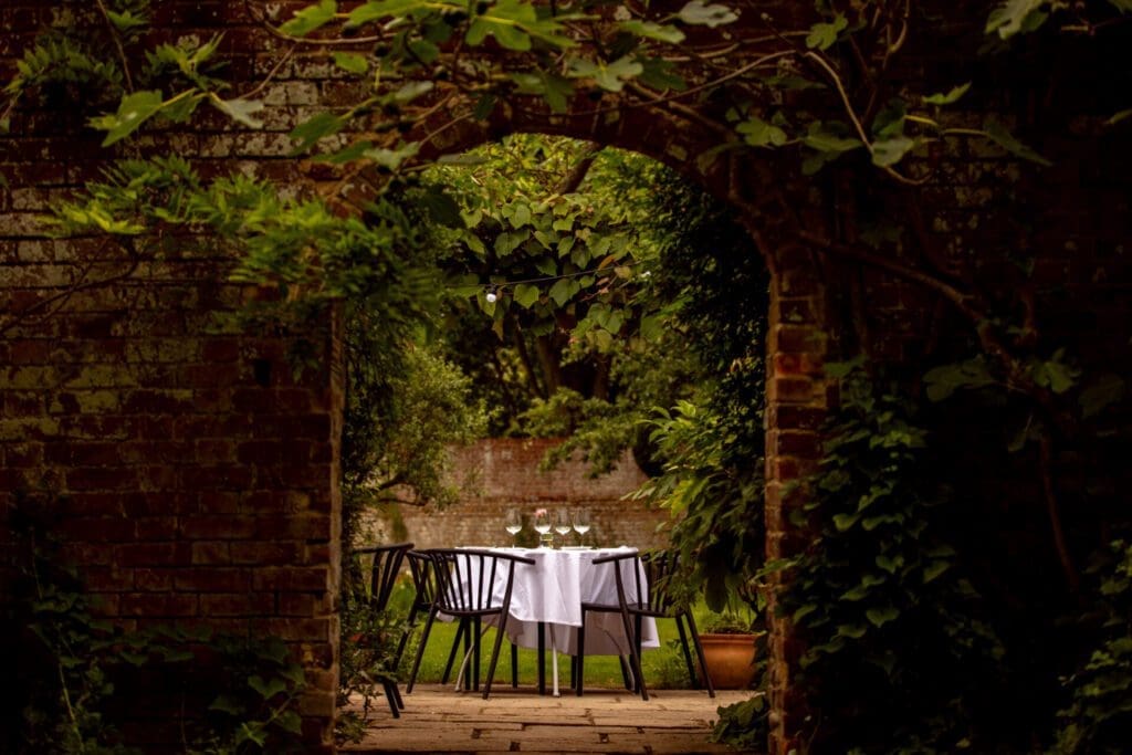 Best new hotels for summer 2022 | A table with two chairs and white table cloth is seen through a gate in a brick wall overgrown with creeping greenery
