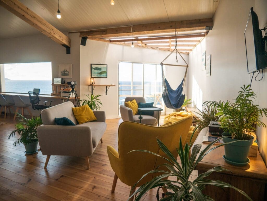 The best European cities for remote working | Inside the lounge and co-working space of Dwell Azores, where sofas and arm chairs and a hammock chair are gathered on the right, and a table against the wall with office chairs shows where to work, with a picture window of the Atlantic.