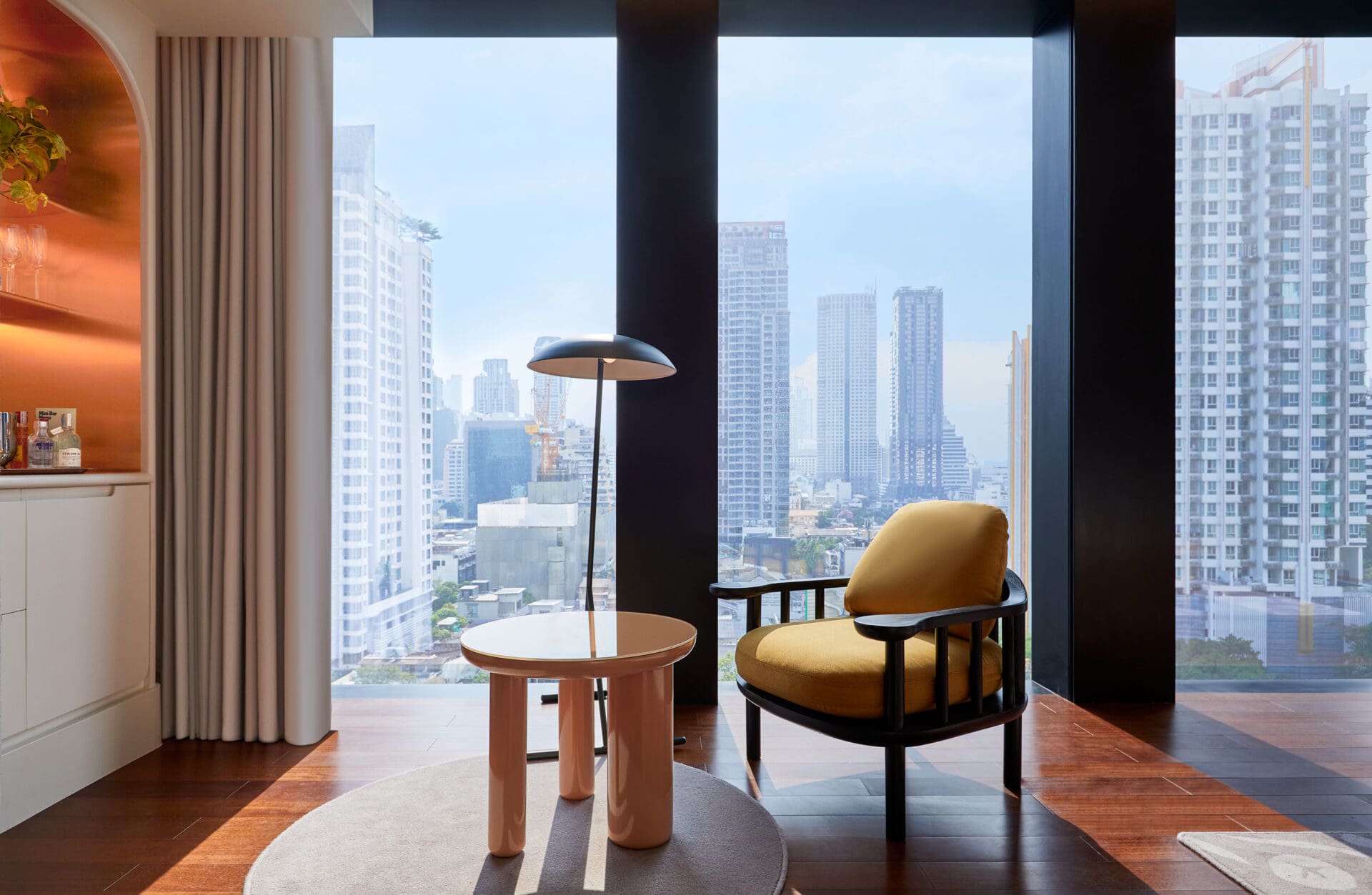Best new hotels for summer 2022 | An armchair with dark wood arms and legs and overstuffed yellow upholstery and a low round table sit in front of floor-to-ceiling windows looking out over a city skyline
