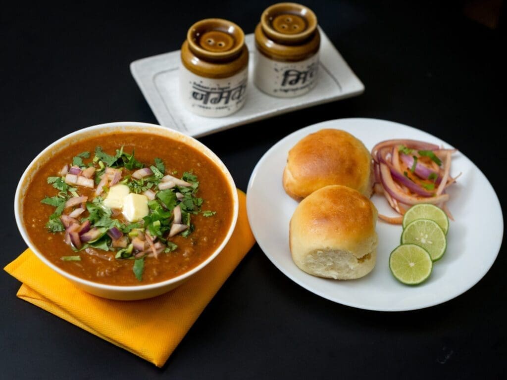 The best street food in Mumbai | pav bhaji: a bowl of tomato curry with fluffy white rolls, limes, and sliced onion on the side.