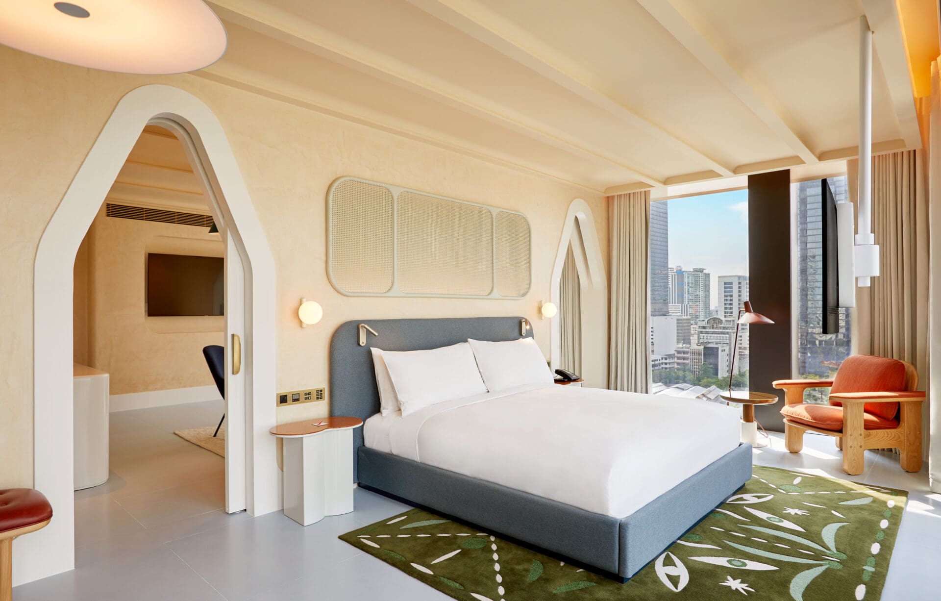 Best new hotels for summer 2022 | a big white bed with blue upholstered headboard sits in a suite at The Standard Bangkok, with a patterned green rug, and an arch-shaped doorway and floor-to-ceiling windows