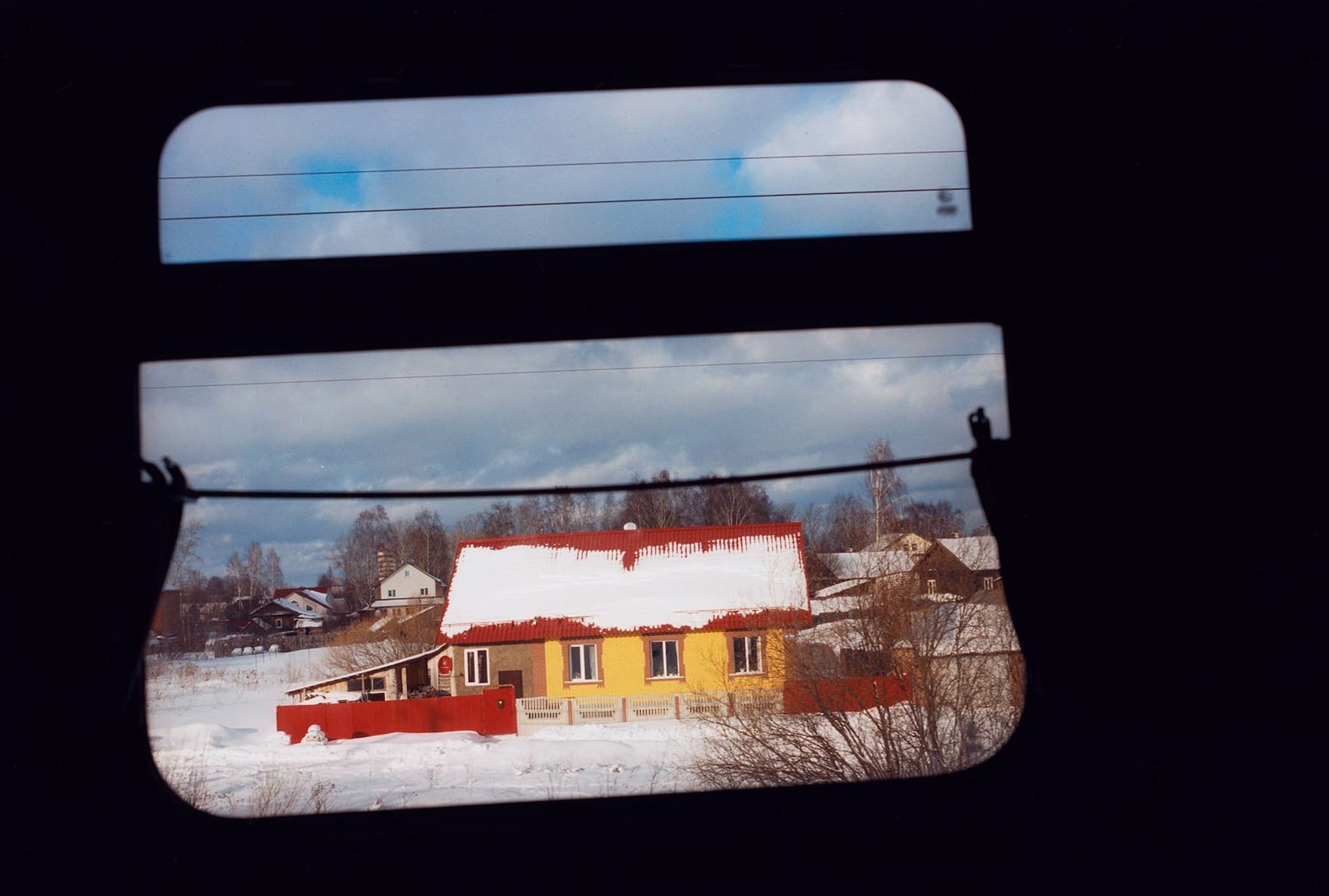 Coco Capitan, Transsiberian | A view through a train window to a yellow house with a red roof covered in snow