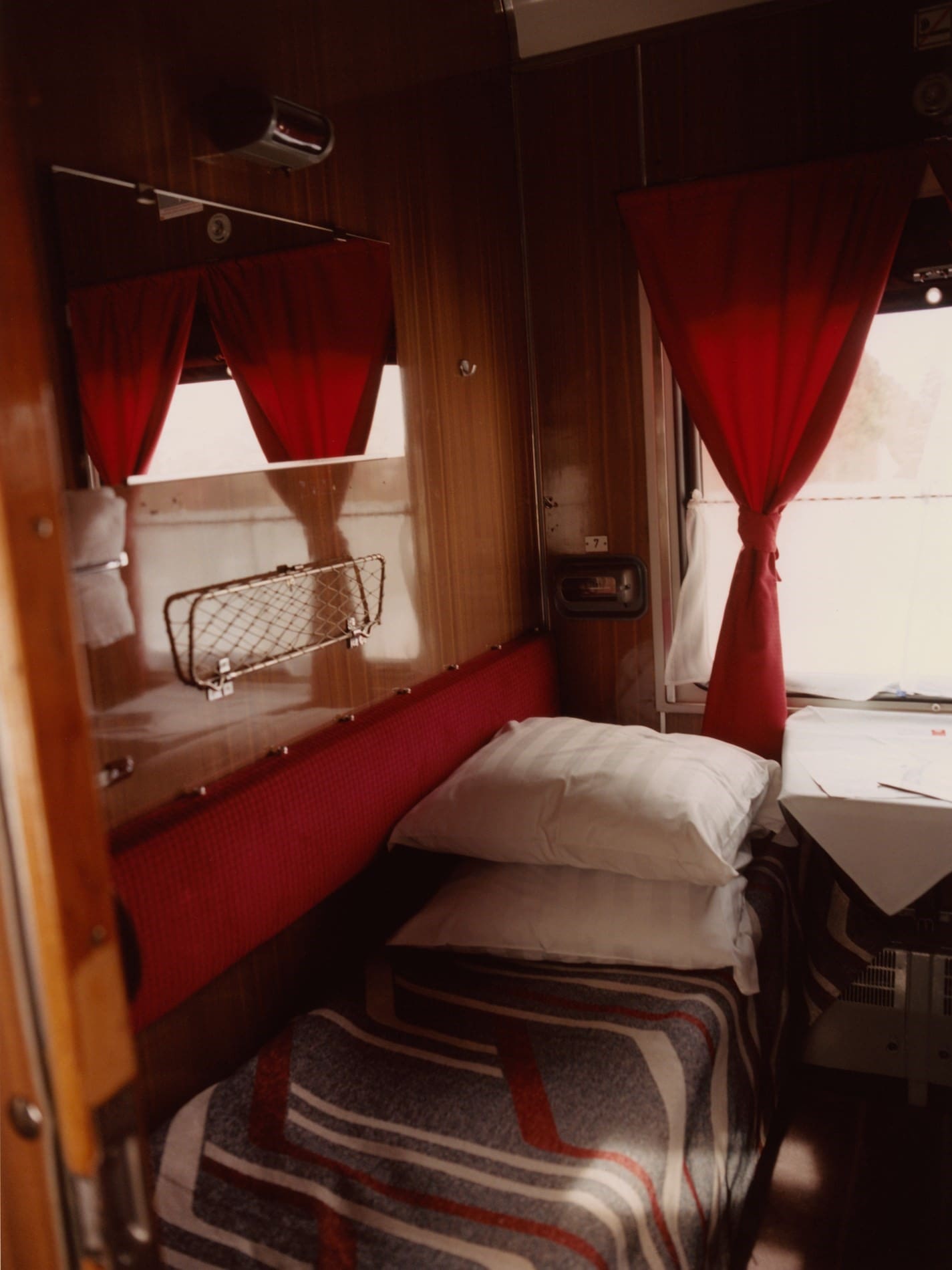 Coco Capitan | A bed inside a train carriage's sleeper compartment, with red curtains, pillows and striped bedding