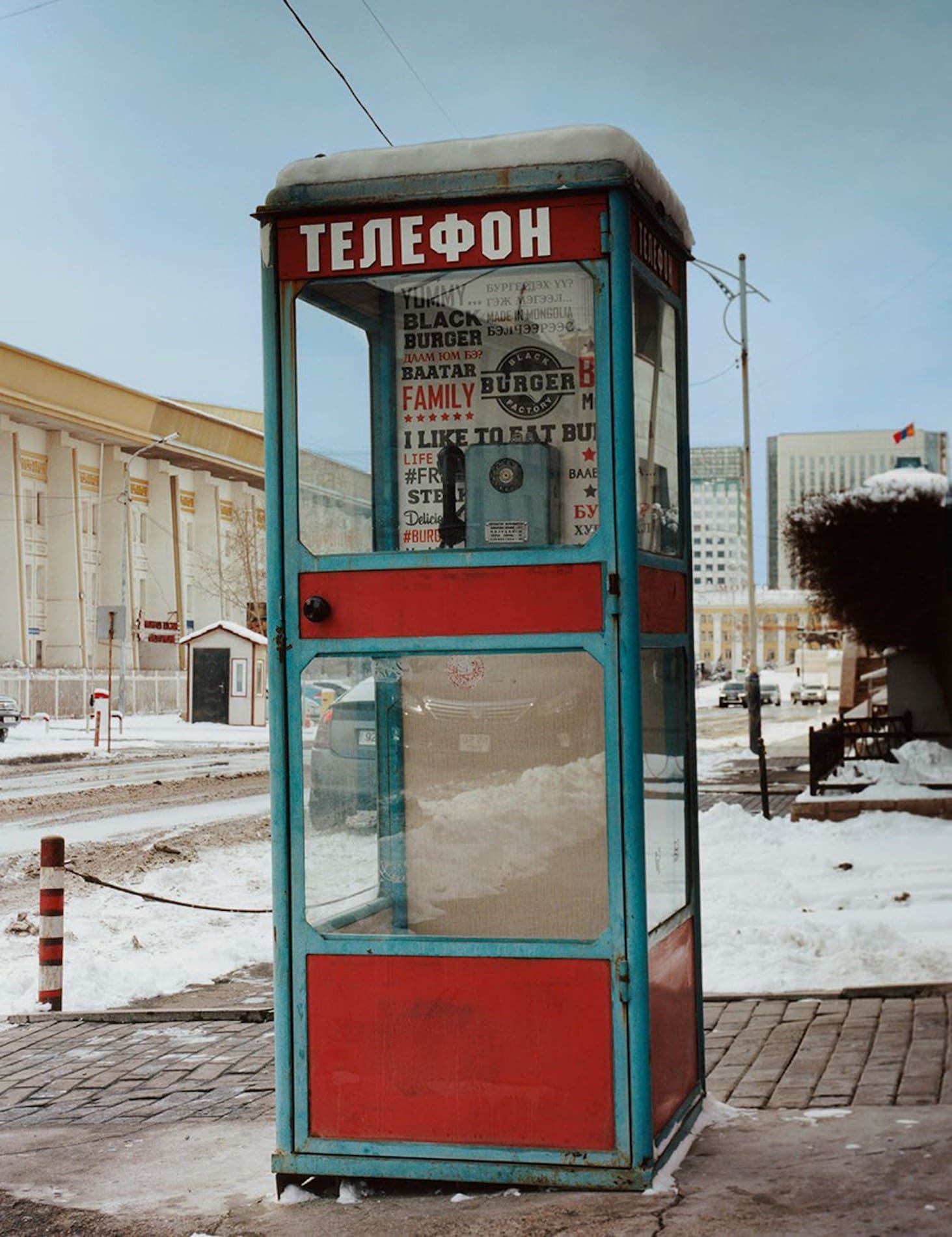 Coco Capitan | A Russian phonebox in Siberia, with red panels and blue frame, on a paved corner by a snow-covered road against a pale-blue sky