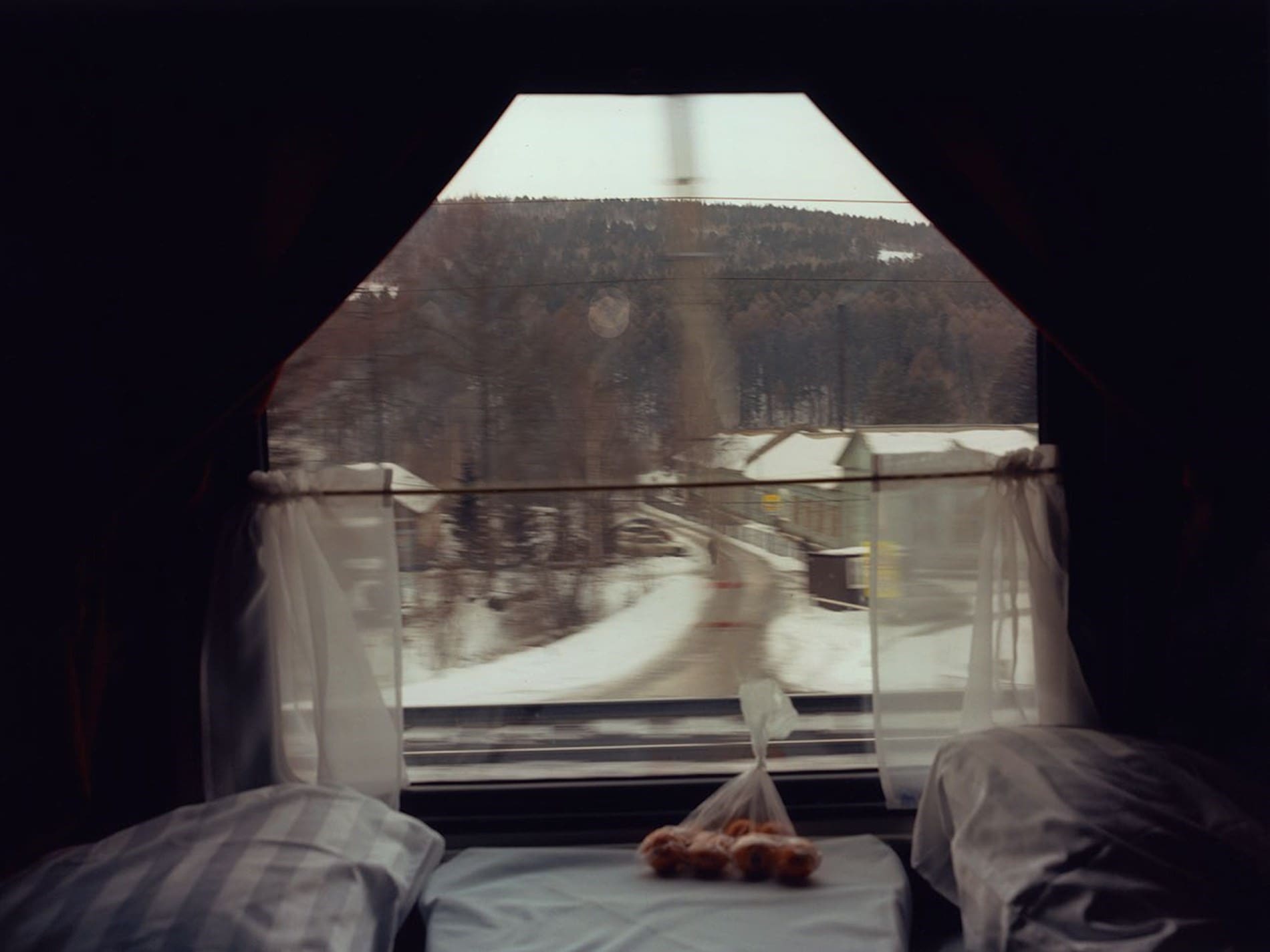 Coco Capitan, Transsiberian | A window by a table seat on a train, with a bag of fruit and looking out at a snow-covered village