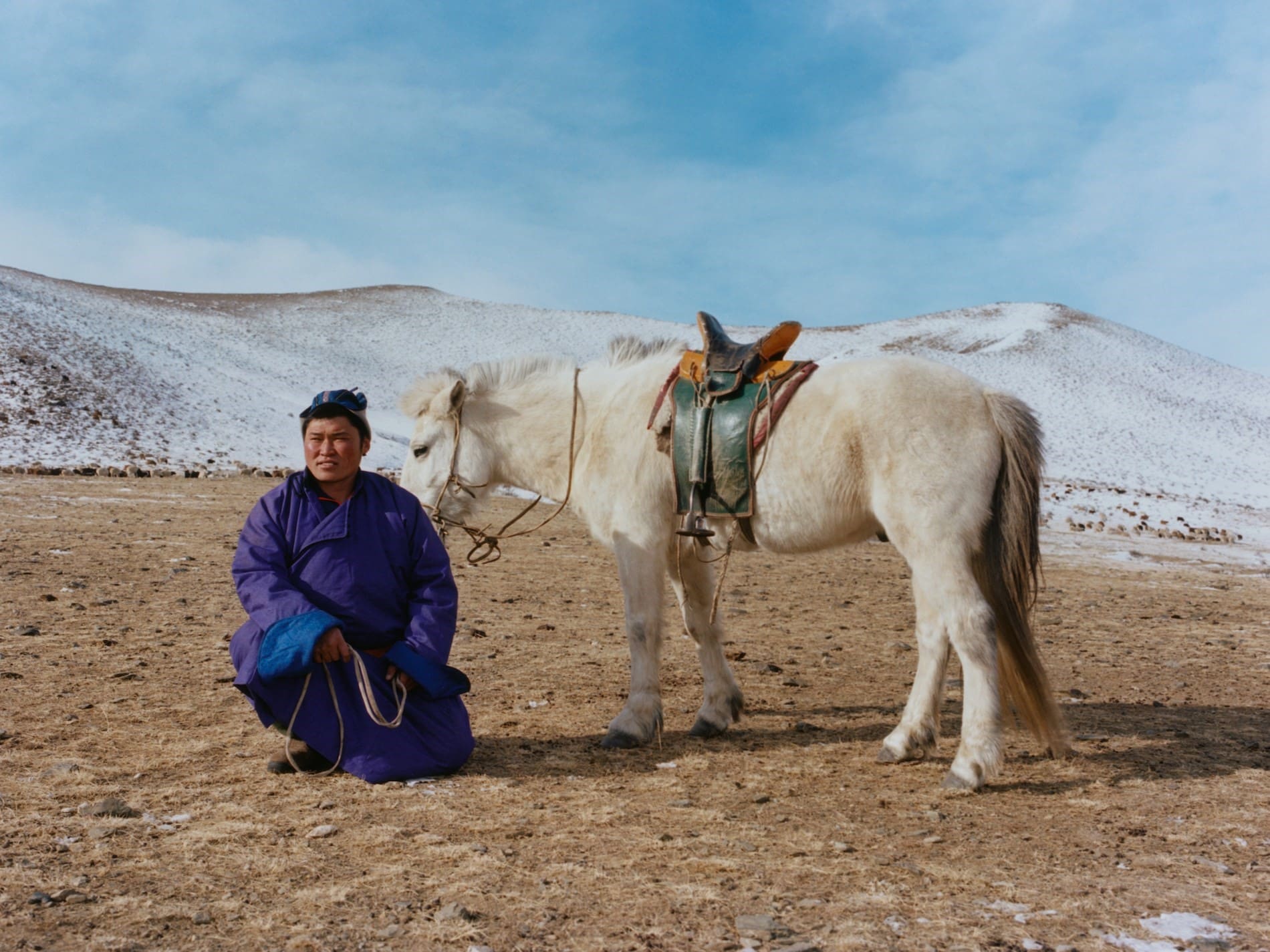 Coco Capitan | A man in a blue and purple outfit sits next to a white pony on earthy terrain, with white mountains and blue sky in the background