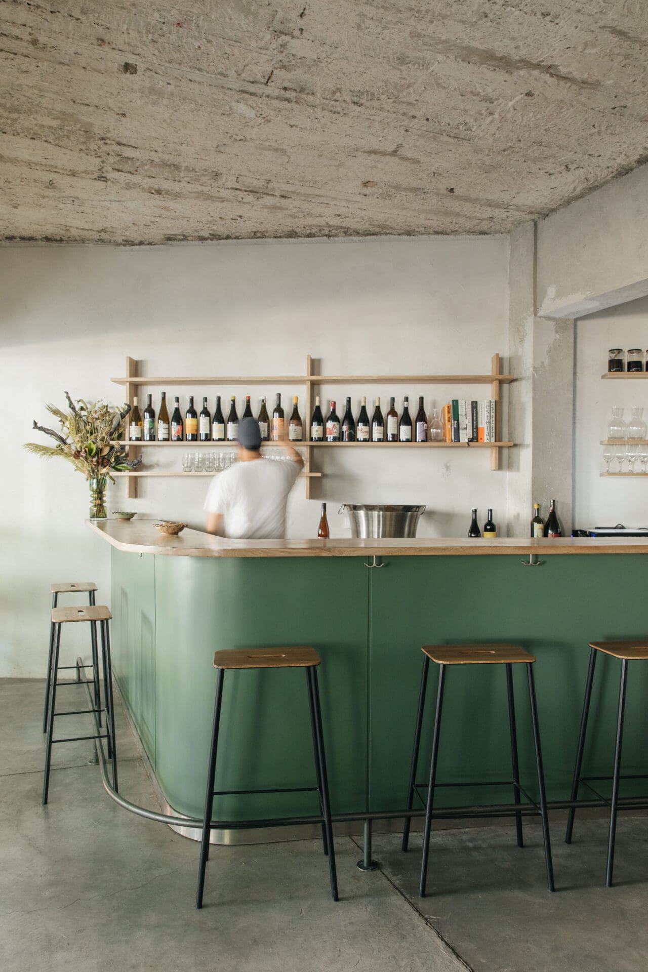 The best bars in Mexico City | A view of the green bar at Hugo El Wine Bar, with stools at the counter and a man reaching for a bottle from a shelf on the white wall