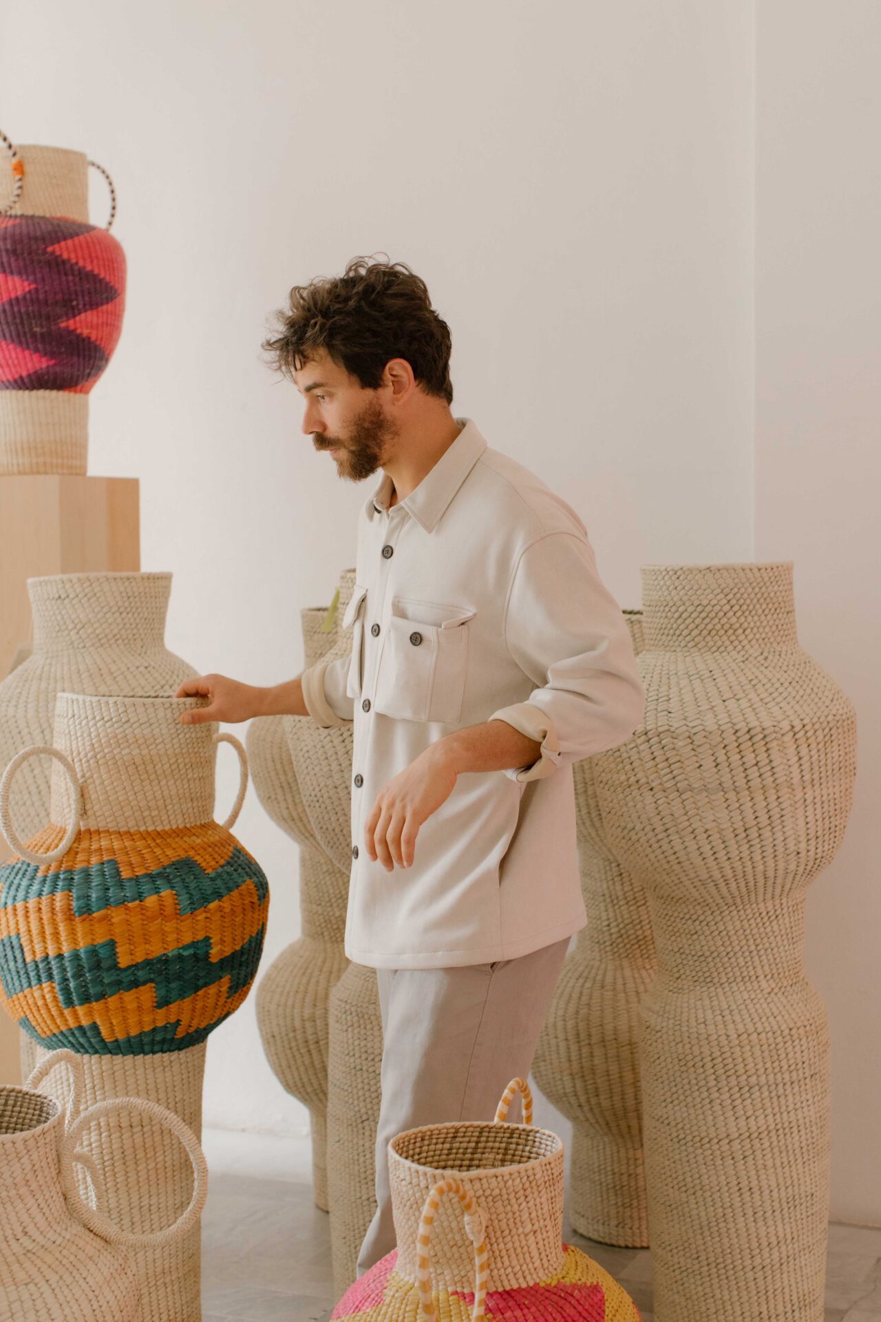Javier Reyes, rrres studio, Oaxacao | the designer in his studio, wearing a white outfit and looking at some colourful woven baskets