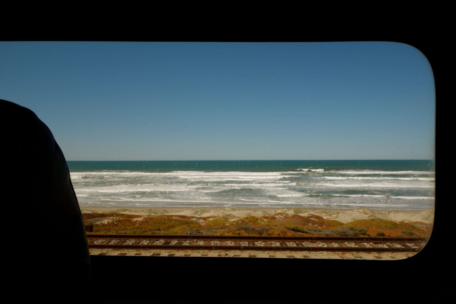 Travelling from LA to San Francisco via Amtrak train | A view out of an oblong-shaped train window of the California coastline, looking over the sea and the train tracks