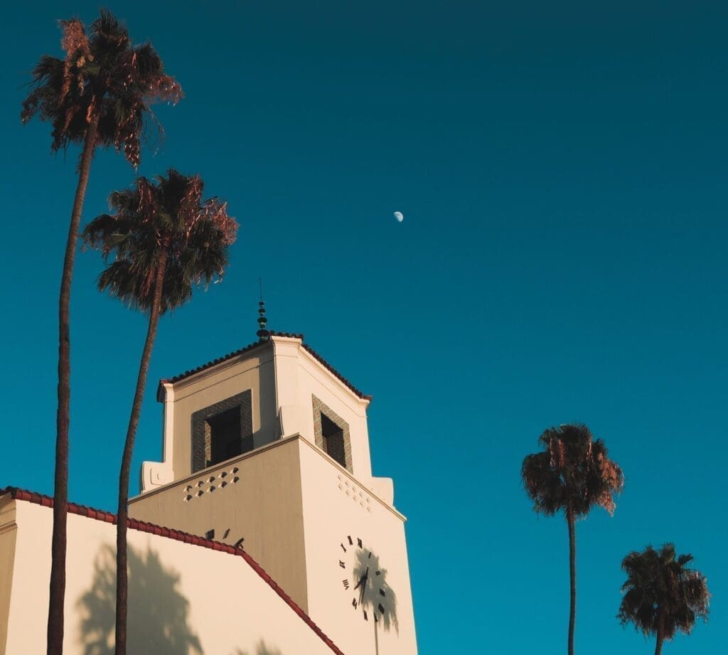 A view of the LA's art deco Union Station, looking up at the clock tower framed by palm trees against a blue sky and a sliver of moon