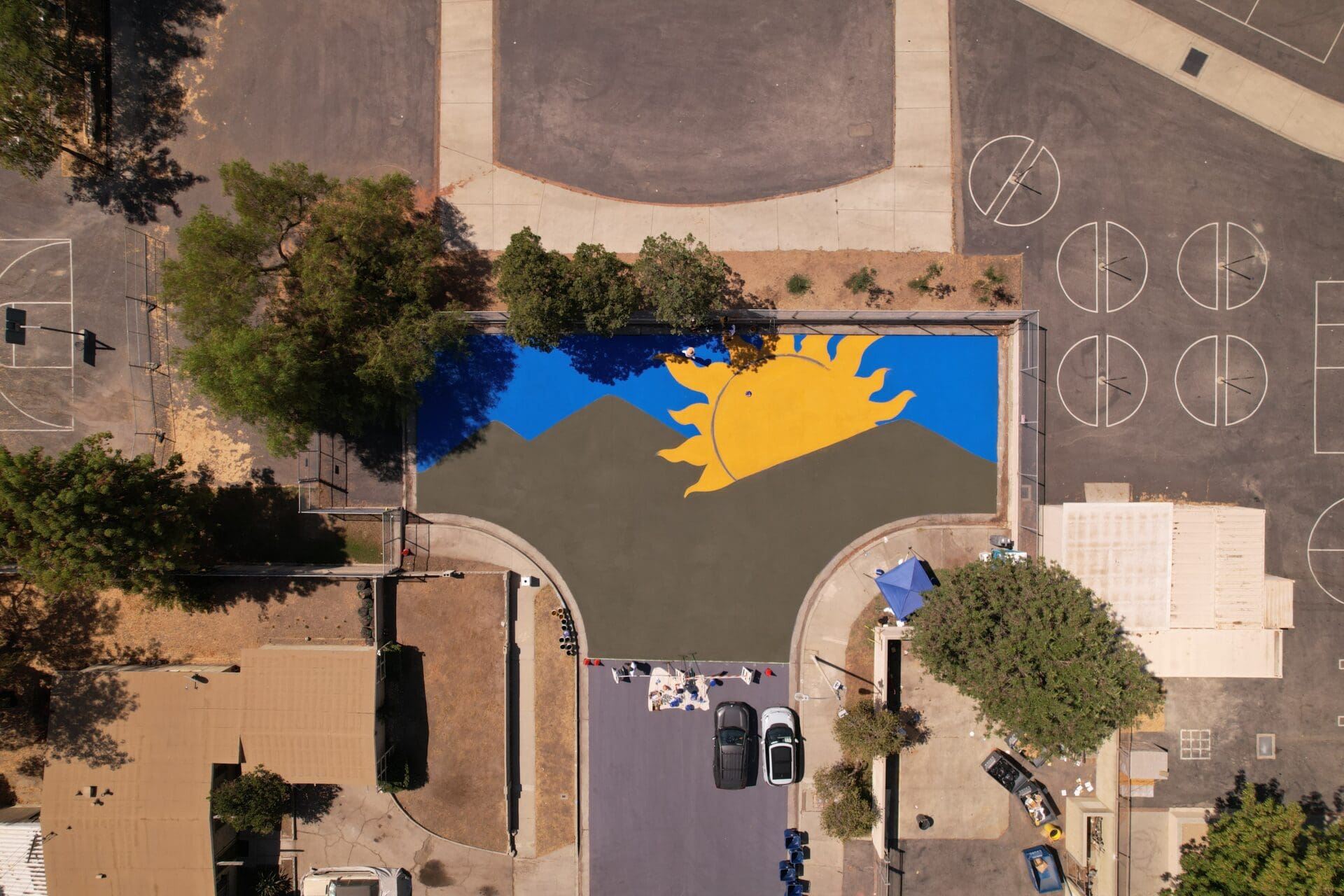 An aerial view of a work-in-progress neighbourhood mural by artist Desiree Sanchez painted on a community park in Pacoima, Los Angeles, featuring a yellow sun and a blue sky