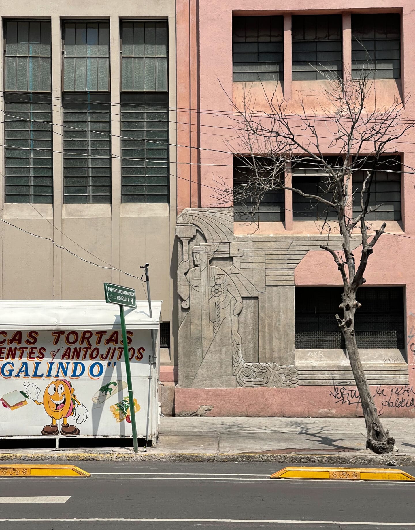 Photographer Manuel Zúñiga on Mexico City | A peach-pink stone building next to a grey stone building, with a bare-branched tree and a small food stall in front of them on a sidewalk