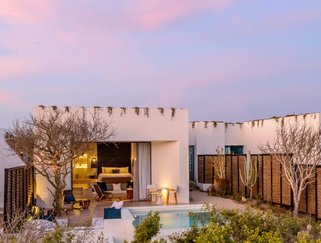 A white open plan villa at Rancho Pescadero, Mexico, framed by cacti beneath a pink-toned sky 