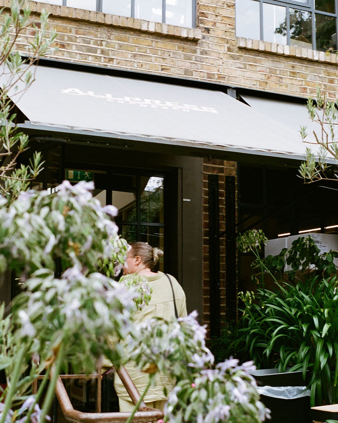 Woman entering Allpress coffee shop in Dalston, surrounded by greenery