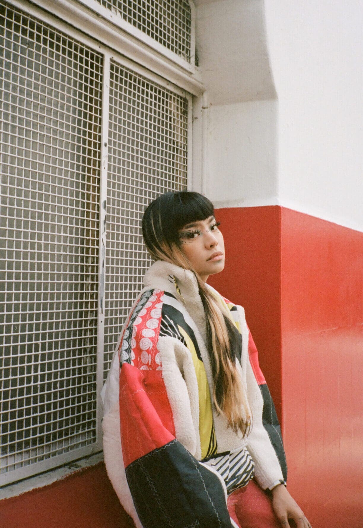 An interview with Thai pop singer Pyra | Pyra sat in front of a metal window grid, leaning against a red and white wall.