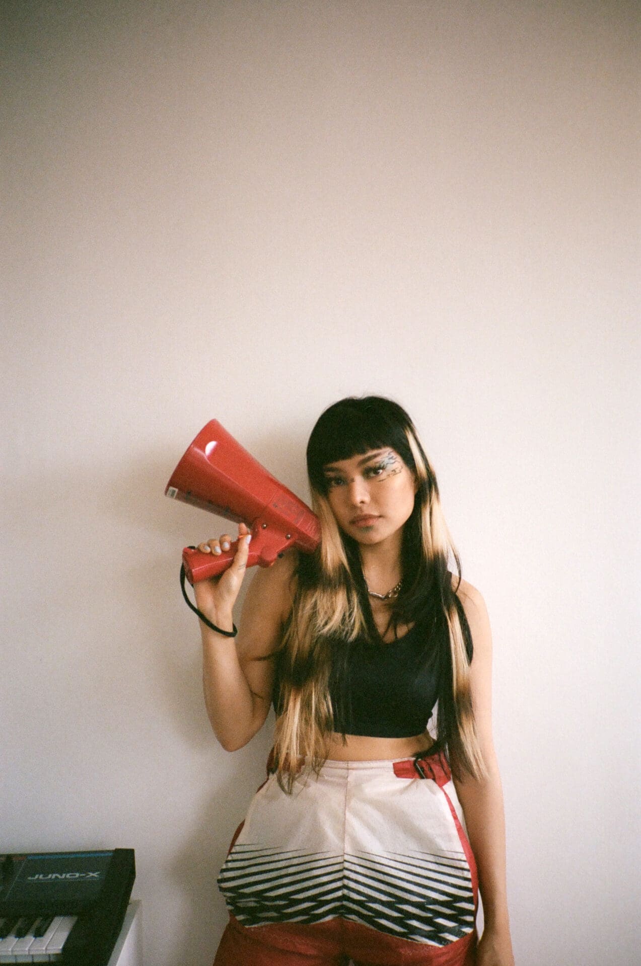 An interview with Thai pop singer Pyra | Pyra standing against a white wall inside, holding a red speakerphone in her right hand, with a Juno synthesiser in the left hand corner of the image.