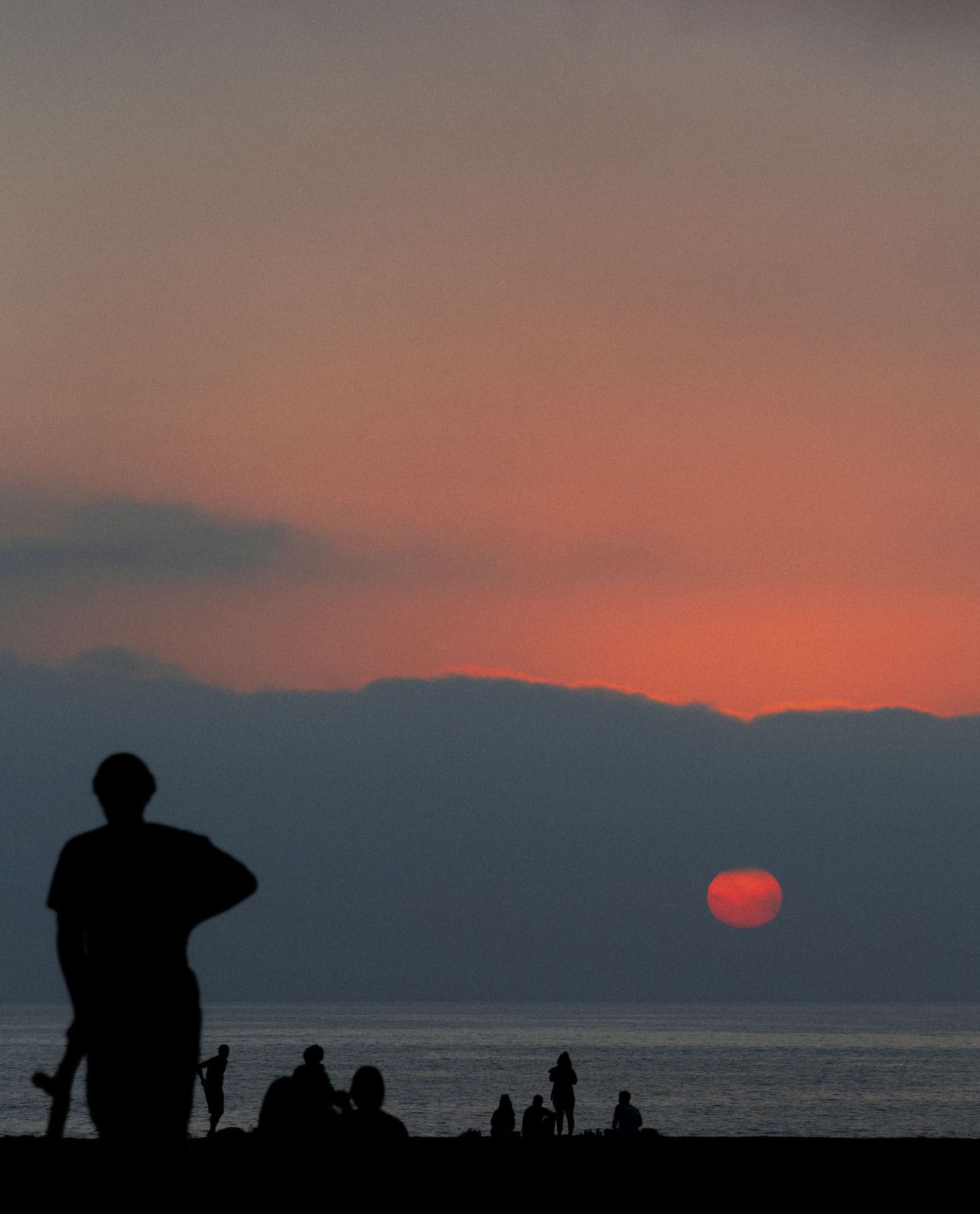 Focal Point, Erik Melvin | Skaters silhouetted against the Pacific Ocean as the red sun dips towards the horizon through a bank of blue grey cloud. Photo by Erik Melvin