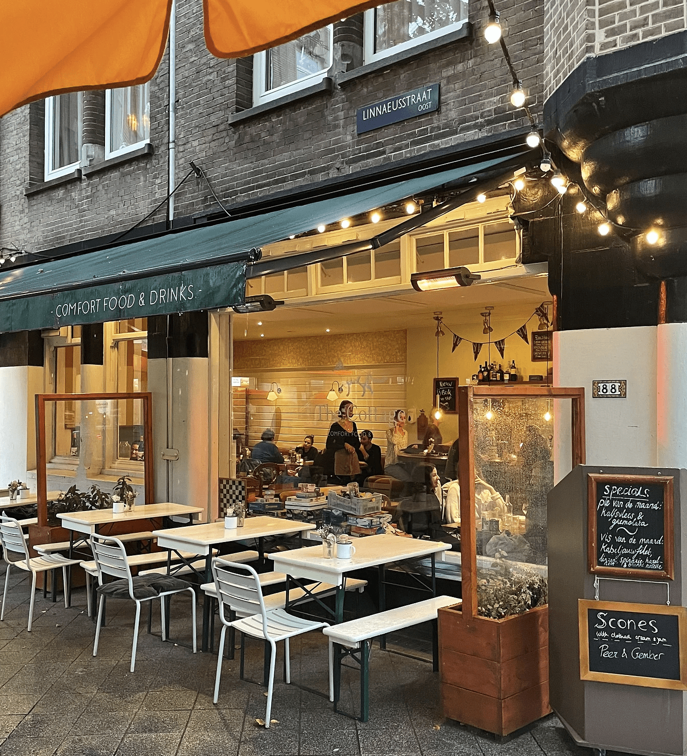 The best restaurants in Amsterdam | The outside of a warm lit restaurant called The Cottage with white tables outside.