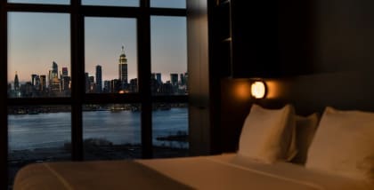A hotel bed by a window at night with NYC skyline views