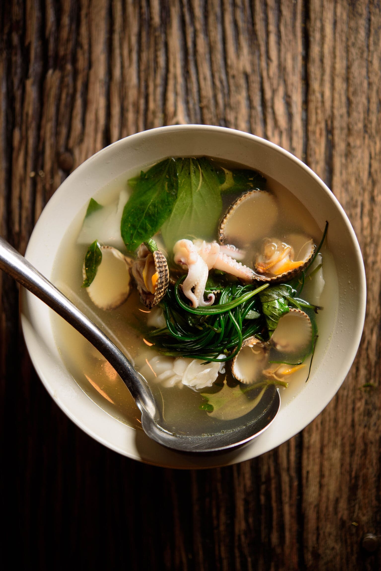 A seafood broth served at Som Saa in a white bowl on a wooden table with a steel spoon placed inside.