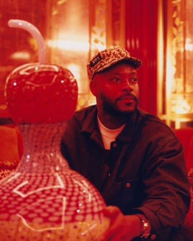 A portrait of Yinka Ilori sat with his original Murano glass sculpture in the red-lit interiors of Mayfair private member's club George