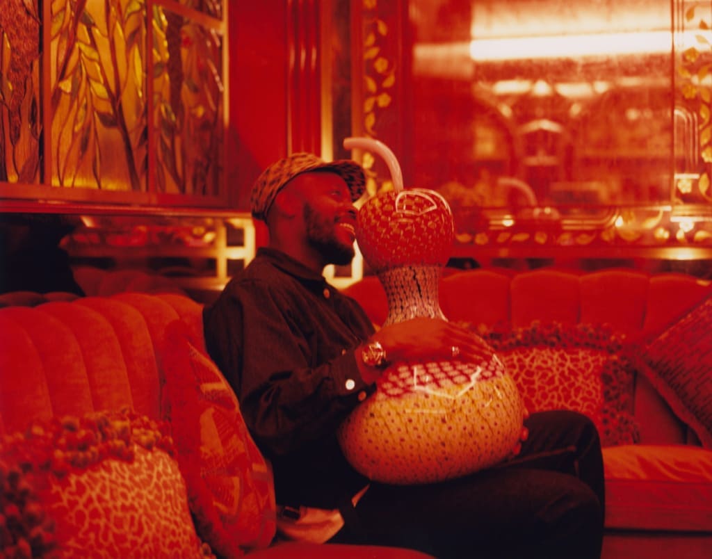 A portrait of Yinka Ilori sat with his original Murano glass sculpture in the red-lit interiors of Mayfair private member's club George.