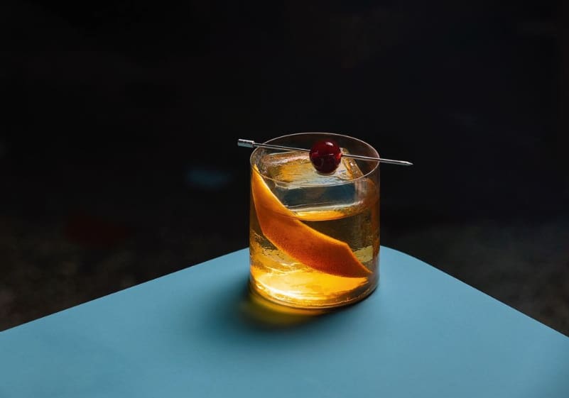 An Old Fashioned with Woodford Reserve bourbon, cherry, and Angostura bitters, served on a blue tabletop at Offtrack cocktail bar.