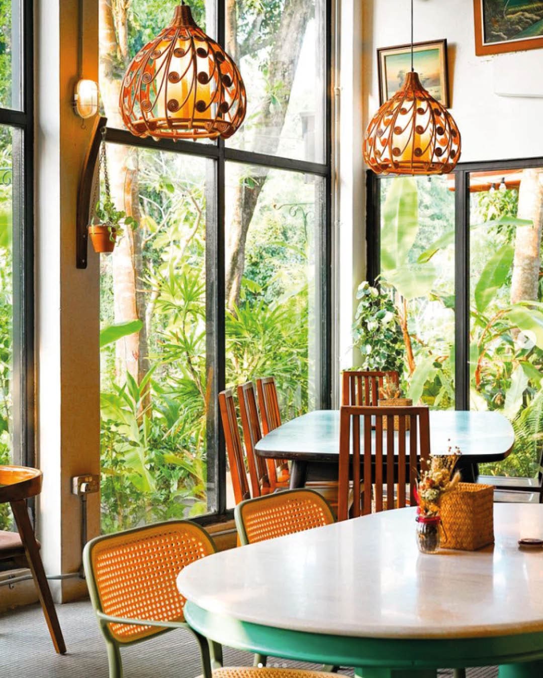 Chairs and tables beneath hanging pendants in front of floor-to-ceiling windows in The Coastal Settlement restaurant, Singapore.
