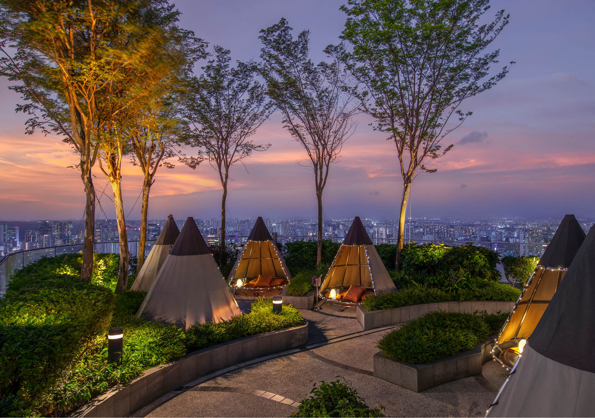 Tipi huts outside Andaz Singapore interspersed with trees, and a view of the city in the evening in the background.